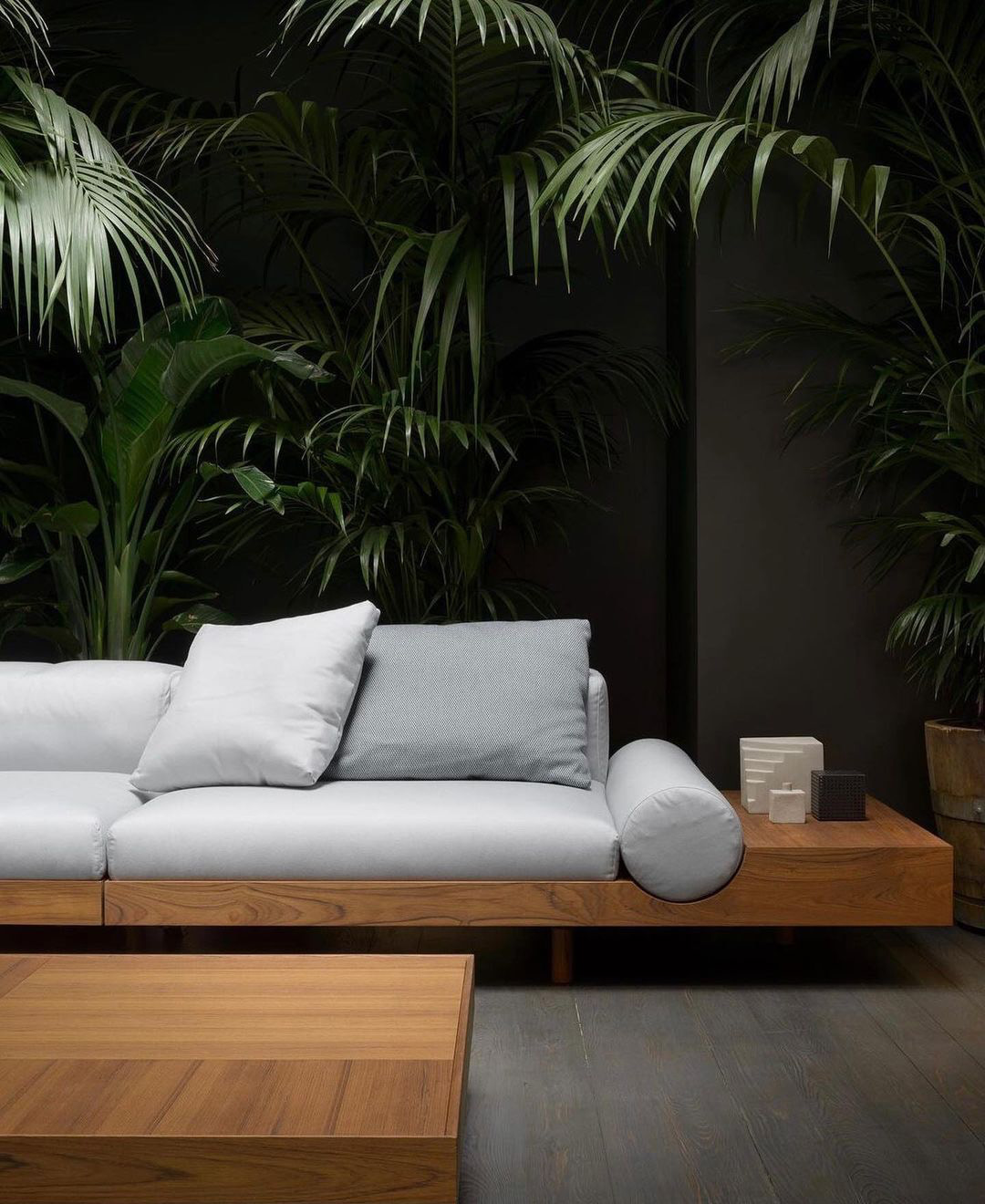 Low timber platform sofa against dark foliage and charcoal plaster wall